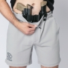 A man wearing light gray FN-branded drawstring shorts demonstrates concealed carry by pulling down the waistband to reveal a pistol and a magazine secured in a black tactical holster belt